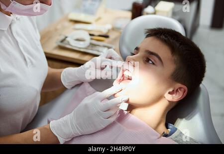 Gros plan d'un enfant garçon confiant avec un enrouleur de joue dans la bouche assis dans la chaise du dentiste, regardant son dentiste traitant pendant le traitement dentaire oral Banque D'Images