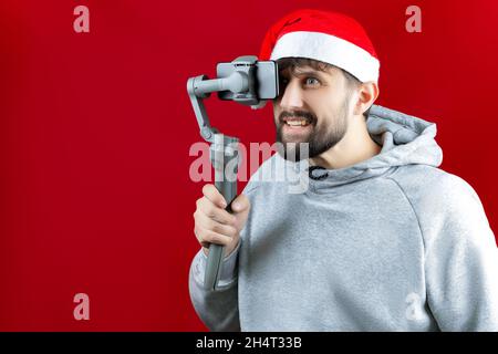 Un homme dans un chapeau de Noël du Père Noël tient un gadget stabilisateur d'image devant lui sur lequel le téléphone est fixé Banque D'Images