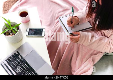 Femme couvrant couverture rose assis sur un canapé prenant des notes sur un ordinateur portable sur une table. Apprendre ou travailler à la maison. Banque D'Images