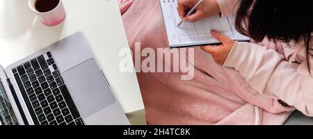 Femme couvrant couverture rose assis sur un canapé prenant des notes sur un ordinateur portable sur une table. Apprendre ou travailler à la maison. Banque D'Images