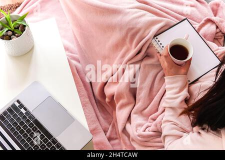 Femme couvrant couverture rose tenant un thé assis sur un canapé avec un carnet . apprendre ou travailler à la maison. Banque D'Images
