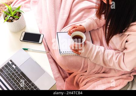 Femme couvrant couverture rose tenant un thé assis sur un canapé avec un carnet . apprendre ou travailler à la maison. Banque D'Images