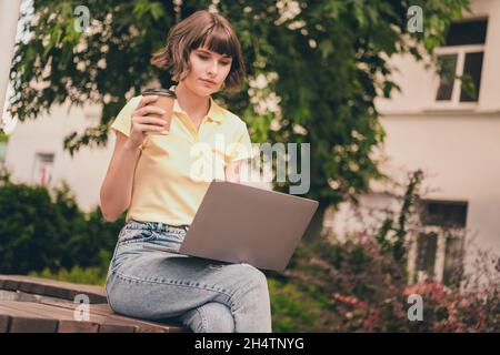 Photo de la stricte femme brunette millénaire travail ordinateur portable boisson thé vêtements t-shirt jeans dehors dans le parc Banque D'Images