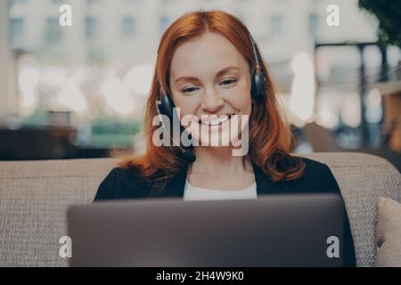 Souriante heureuse jeune femme à tête rouge portant un micro-casque parlant avec des collègues en ligne sur un ordinateur portable Banque D'Images