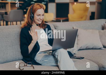 Jeune femme utilisant un ordinateur portable et un casque pour les appels vidéo sur Internet en agitant la main dans le geste de salutation Banque D'Images
