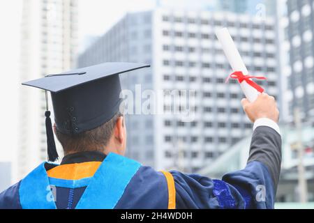 Bon diplômé.Heureux Asain homme dans les robes de graduation tenant un diplôme en main sur fond de ville urbaine. Banque D'Images