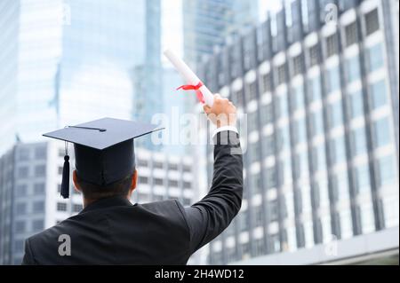 Bon diplômé.Heureux Asain homme dans les robes de graduation tenant un diplôme en main sur fond de ville urbaine. Banque D'Images