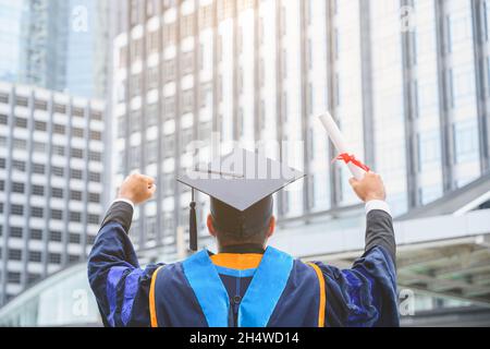 Bon diplômé.Heureux Asain homme dans les robes de graduation tenant un diplôme en main sur fond de ville urbaine. Banque D'Images