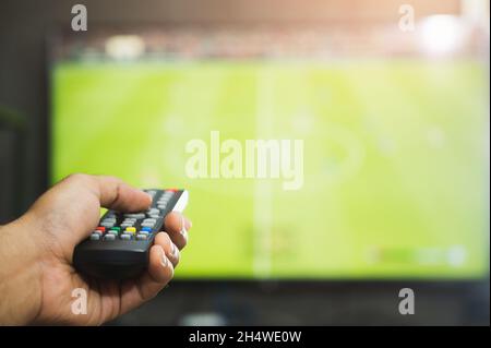 Jeune homme tenant la télécommande de télévision regardant un programme de football.Mains pointant vers le téléviseur et l'allumer ou l'éteindre pour sélectionner le canal de surveillance Banque D'Images