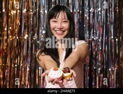 Une jeune femme asiatique montre des cupcakes à l'appareil photo sur un fond de fête. Banque D'Images