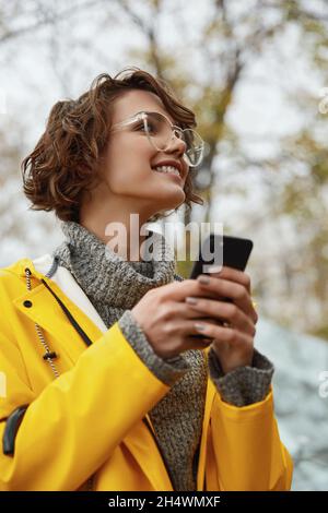 Jeune fille en imperméable jaune avec smartphone à la main marchant dans la ville.Jeune belle femme brune aux cheveux vêtue d'un vêtement de pluie tout en allant sur la rade Banque D'Images