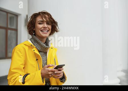 Jeune fille en imperméable jaune avec smartphone à la main marchant dans la ville.Jeune belle femme brune aux cheveux vêtue d'un vêtement de pluie tout en allant sur la rade Banque D'Images