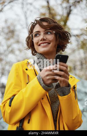 Jeune fille en imperméable jaune avec smartphone à la main marchant dans la ville.Jeune belle femme brune aux cheveux vêtue d'un vêtement de pluie tout en allant sur la rade Banque D'Images