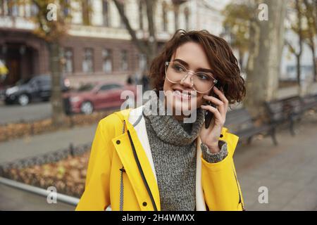 Jeune fille en imperméable jaune qui appelle sur un smartphone tout en marchant dans la ville.Jeune belle femme brune aux cheveux vêtue de vêtements de pluie pendant que vous allez Banque D'Images