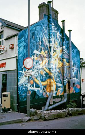 «Fishing», une fresque d’Edward Povey sur le côté d’un magasin de poissons et de puces situé au 27-29 High St, Menai Bridge Anglesey, pays de Galles, en 1987.La peinture murale a été commandée par le propriétaire de l’atelier de l’époque, M. Radford (senior), et a été peinte à base d’huile sur le rendu de ciment apprêté en juin 1980.Il représente un chalutier de pêche et des pêcheurs en mer.Il est toujours en place, mais il est défraîchi et endommagé par le temps – le bâtiment est maintenant un restaurant indien.Artiste anglais, Edward Povey (n.1951), peint 25 peintures murales sur des bâtiments du pays de Galles et du nord-ouest de l'Angleterre entre 1975 et 1985. Banque D'Images