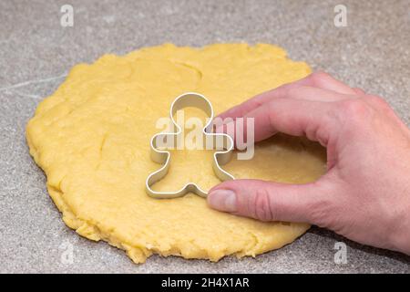 La main du cuisinier coupant la forme des hommes de pain d'épice de pâte roulée crue sur un fond gris.Le processus de fabrication de biscuits de Noël à h Banque D'Images