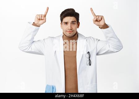 Un médecin confus pointant des doigts vers le haut, regardant perplexe, debout dans une robe médicale sur fond blanc Banque D'Images