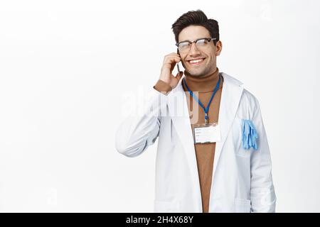 Un médecin souriant répond à un appel téléphonique, parle au téléphone portable, porte un peignoir d'uniforme de clinique, debout sur fond blanc Banque D'Images