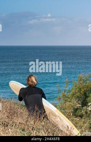 Un homme enthousiaste surfeur transportant sa planche de surf marchant sur un sentier côtier vers la mer dans les Cornouailles. Banque D'Images