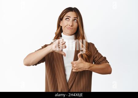 Indécis femme d'affaires d'âge moyen pesant la décision, montrant comme un geste déplaît, pouces vers le haut et pouces vers le bas, regardant vers le haut et pensant, blanc Banque D'Images