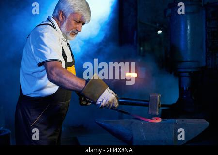 Forgeron caucasien expérimenté dans le tablier de protection et les gants de cintrage de l'acier sur l'enclume.Travailleur industriel mature utilisant des pinces et un marteau à forge. Banque D'Images