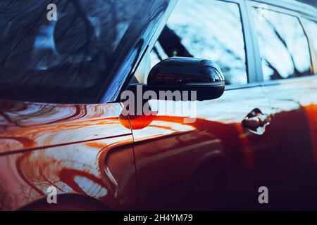 Une voiture neuve rouge, chère et polie, descend la rue, reflétant le ciel bleu et les branches des arbres.Transport. Banque D'Images