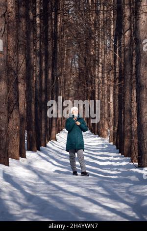 Une jeune femme marche dans une forêt enneigée.La jeune femme en veste de duvet verte et chapeau blanc aime marcher le long du sentier d'hiver parmi les grands arbres Banque D'Images