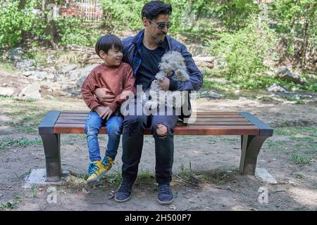 Père et fils latino-américains, et un chiot blanc en peluche, assis sur un banc de bois, reposant après une promenade le long de la crique. Horizontal.Gamme vac Banque D'Images