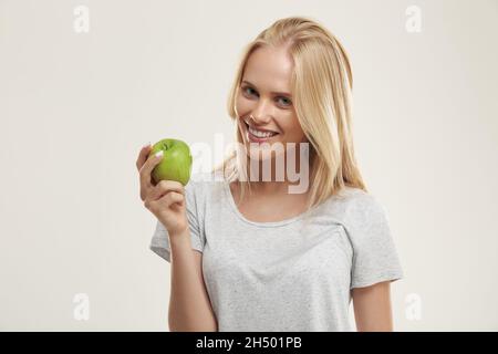 Jeune fille européenne souriante tenant une pomme verte.Belle femme blonde adolescente avec les yeux bleus porter un t-shirt blanc et regarder l'appareil photo.Isolé sur Banque D'Images