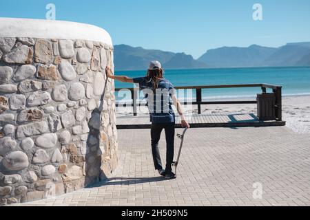 Vue arrière d'un homme tenant une planche à roulettes penchée sur le mur de la promenade tout en regardant la mer Banque D'Images