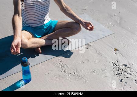 Bas section de l'homme méditant en pratiquant le yoga par bouteille d'eau sur le sable à la plage Banque D'Images