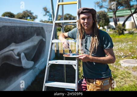 Portrait d'un artiste masculin tenant un pinceau et une palette penchée sur une échelle par une murale représentant une baleine Banque D'Images