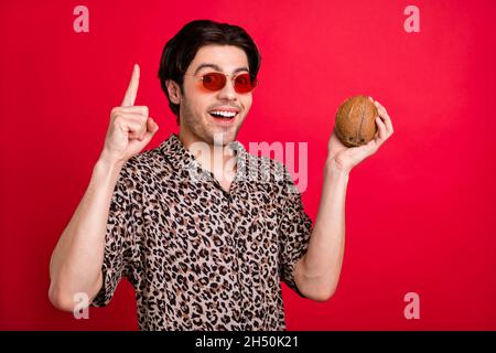 Photo de sourire gai beau homme tenir le point de noix de coco copyspace recommander des conseils isolés sur fond rouge couleur Banque D'Images