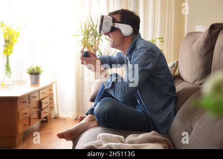 Amusant homme jouant avec des lunettes de réalité virtuelle avec un joystick dans ses mains assis sur un canapé à la maison Banque D'Images