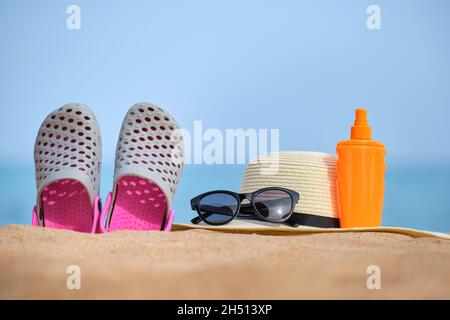 Gros plan sur un chapeau de paille, des chaussures de sabots, de la crème solaire et des lunettes de soleil de protection noires sur une plage de sable au bord de la mer tropicale par beau temps ensoleillé. Vacances d'été con Banque D'Images