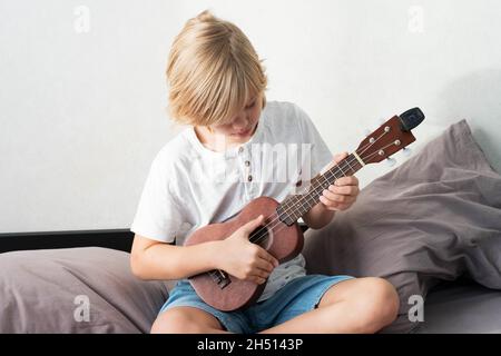 Jeune garçon tuning ukulele à la maison.Garçon blond aux cheveux assis sur un canapé jouant de la guitare acoustique. Banque D'Images