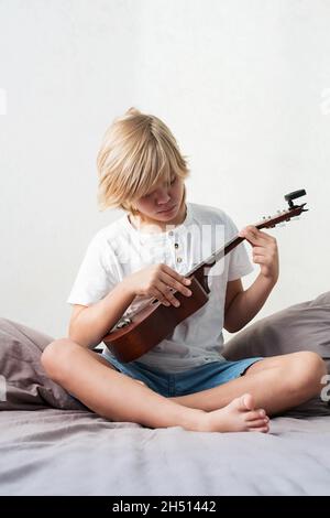 Jeune garçon tuning ukulele à la maison.Garçon blond aux cheveux assis sur un canapé jouant de la guitare acoustique. Banque D'Images