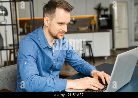 Professionnel indépendant sérieux et intelligent utilisant un ordinateur portable pour travailler en ligne, assis à son bureau dans l'espace de travail, un homme attentif tapant sur le clavier, écrit des articles, bavarder et envoyer des messages en ligne Banque D'Images