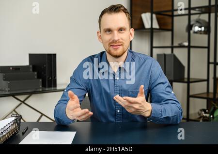 Portrait d'un jeune barbu attrayant parlant à l'appareil photo, assis dans le bureau moderne, portant une chemise décontractée.Concept d'appel vidéo, visioconférence avec un collègue de sexe masculin Banque D'Images