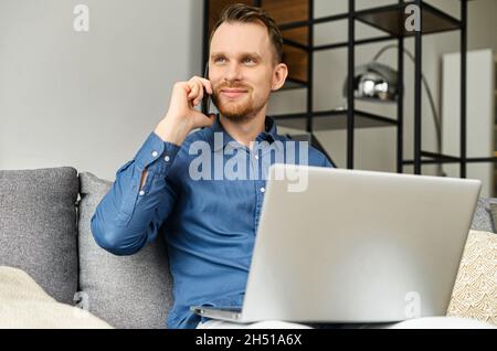 Homme d'affaires sympathique parlant sur le smartphone assis sur le canapé avec un ordinateur portable sur ses tours, un homme indépendant travaillant à distance de la maison, utilisant un ordinateur portable et tenant la conversation téléphonique Banque D'Images