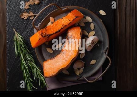 Vue à plat sur les tranches de citrouille cuites sur un plateau en métal sur une table en bois rustique de cuisine avec gousses d'ail et romarin Banque D'Images