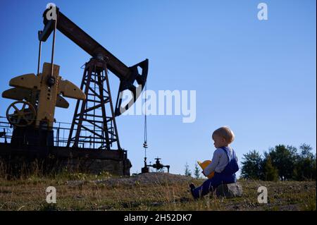 Vue arrière d'un petit garçon d'âge préscolaire assis sur une pierre, tenant le casque de construction dans les mains.Enfant qui cherche à exploiter une station de forage pour l'extraction de pétrole à l'extérieur sous un ciel bleu. Banque D'Images