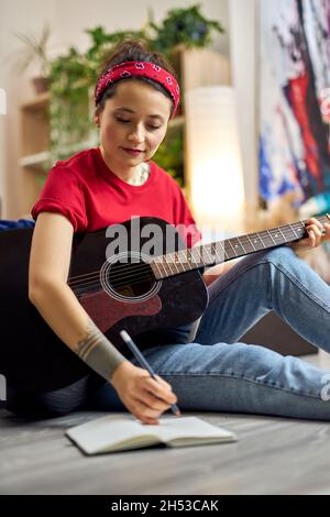 Jeune femme créative en tenue décontractée prenant des notes dans son ordinateur portable tout en jouant de la guitare acoustique, étudiant de la musique à la maison Banque D'Images