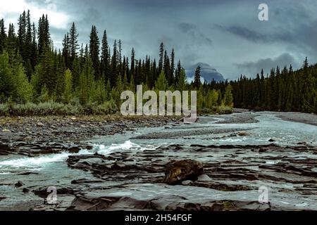 Journée d'été tumultueuse dans les montagnes rocheuses, rivière fluide, arbres lumineux, ciel nuageux sombre, bancs couverts de rochers Banque D'Images