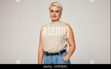 Femme blonde élégante regardant l'appareil photo tout en se tenant sur fond gris.Femme mi-adulte sûre d'elle-même portant un Jean denim dans un studio. Banque D'Images