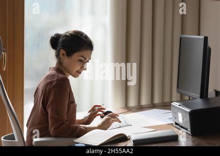 Une femme indienne qui étudie assise au bureau vérifie les informations dans le bloc-notes Banque D'Images
