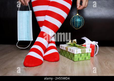 Femme en chaussettes de genou de Noël assis avec masque médical et balle de jouet dans les mains, boîtes-cadeaux sur un plancher.Célébration du nouvel an pendant le coronavirus Banque D'Images