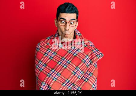 Beau homme hispanique enveloppé dans une couverture rouge chaud faisant le visage de poisson avec la bouche et les yeux accroupidés, fou et comique. Banque D'Images