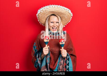 Belle femme blonde caucasienne portant un poncho mexicain festif et des maracas souriant et riant fort parce que drôle de blague folle. Banque D'Images