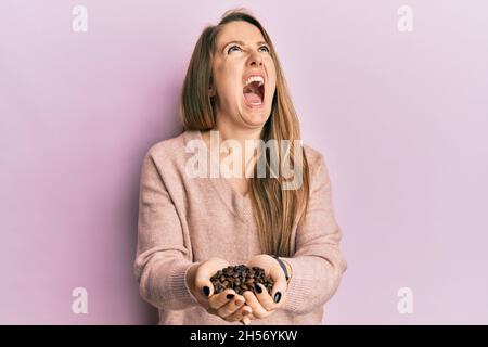 Jeune femme blonde tenant des grains de café avec les mains en colère et folle hurlant frustré et furieux, criant avec colère regardant vers le haut. Banque D'Images